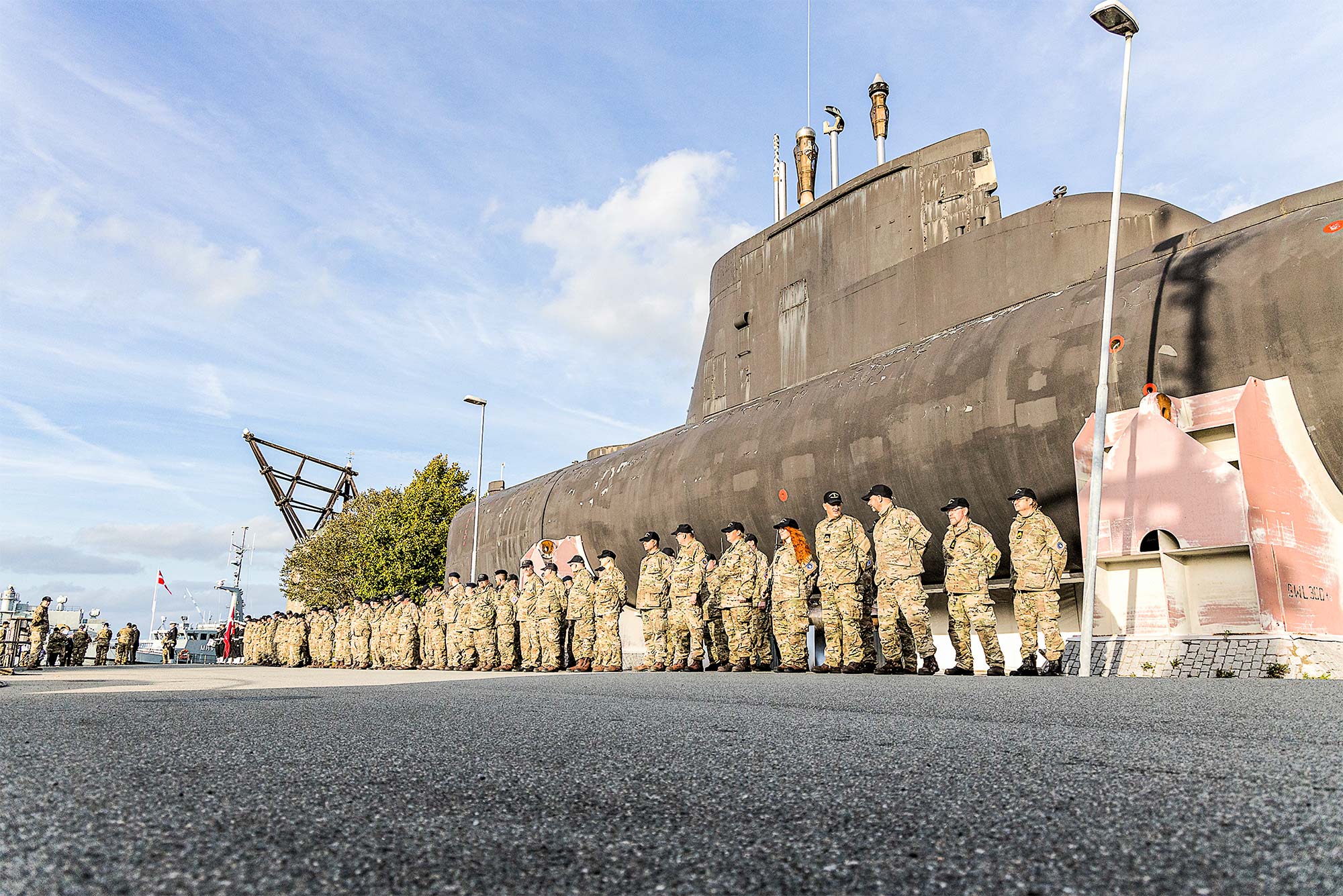 Frivillige marinere samlet til parade på Holmen foran museums-ubåden, Sælen, og den gamle mastekran og rigets flag bagerst. Foto: Andreas Birkebæk.
